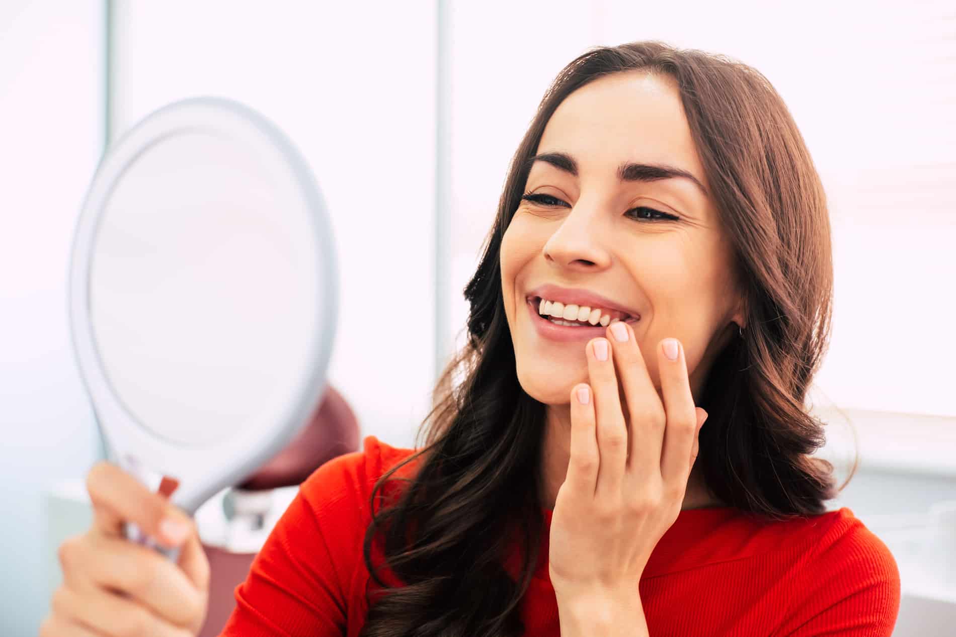 Honed to perfection. Fabulous woman clothed in red sweater, with curly long hair and elegant hands is holding a mirror for looking through the final work of dental doctor. Woman looking at her clean teeth after general dental care with Dr. Chung