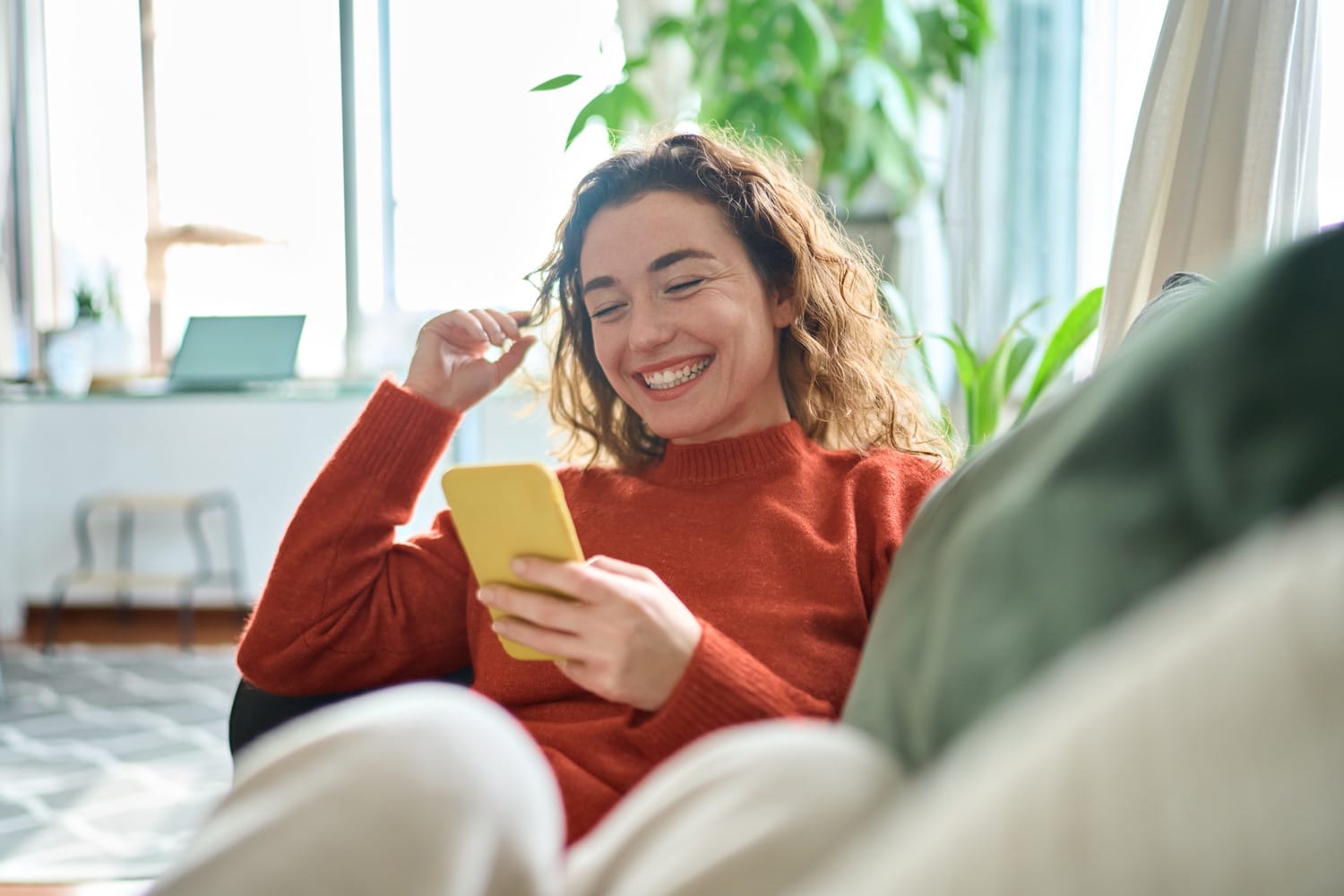 Young woman laughing on the sofa while scheduling a composite bonding dental appointment on her phone in Lindenhurst